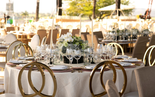 Mesa elegantemente decorada para un evento al aire libre, con centro de mesa floral, copas de cristal y sillas doradas.
