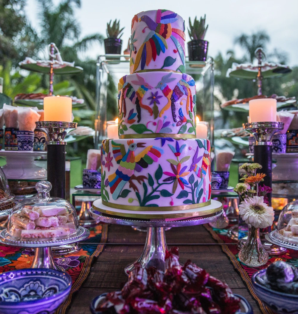Pastel de tres niveles decorado con diseño de bordado otomí, rodeado de dulces mexicanos y velas, en un evento al aire libre.