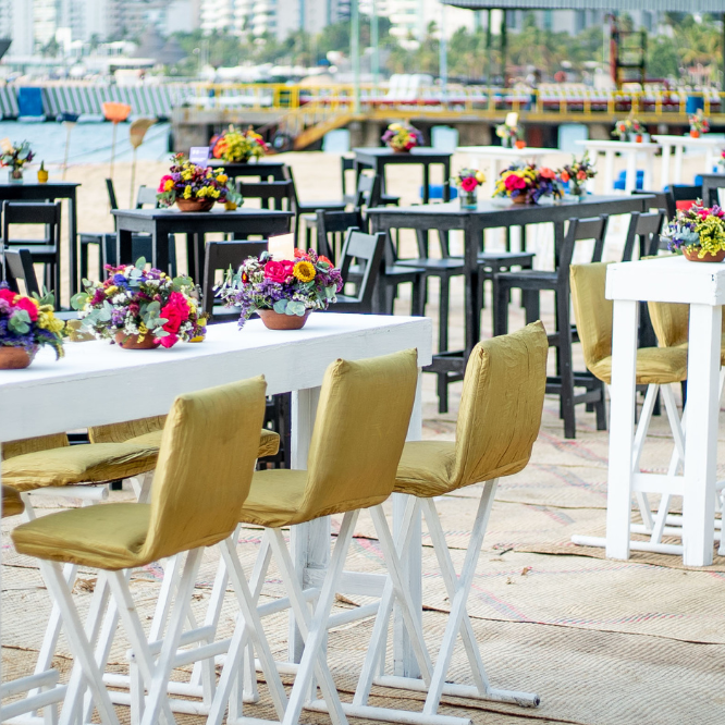 Mesas altas decoradas con flores para un evento en la playa con vista al mar.